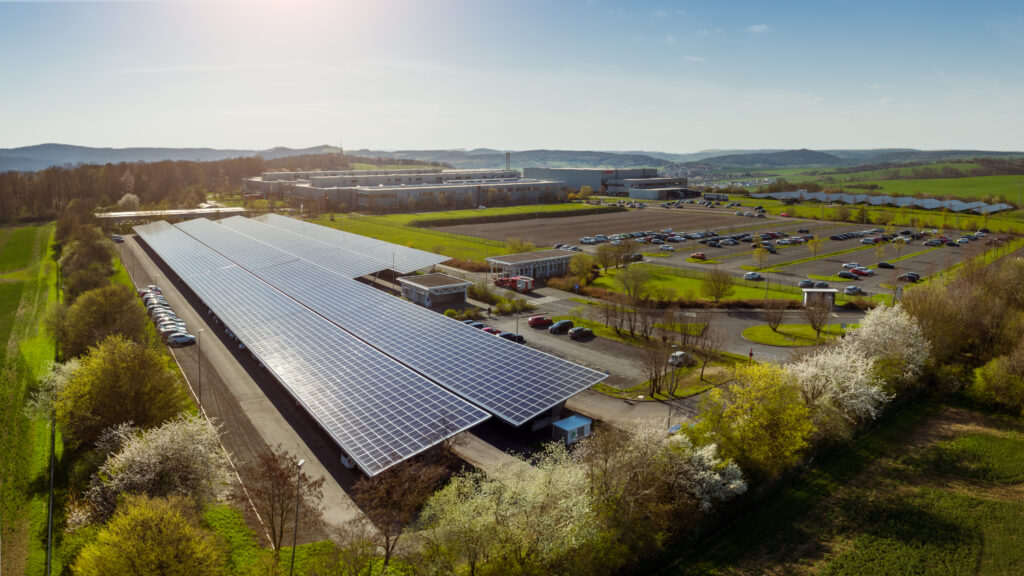 Luftbildaufnahme eines Produktionsstandortes umgeben von grünen Wiesen. Im Vordergrund ein Photovoltaikcarport